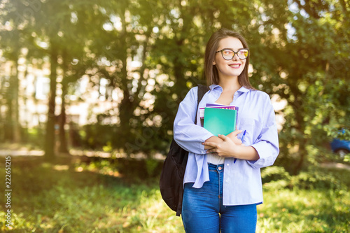 Cheerful attractive young woman student with book in hands backpack standing and smiling in campus