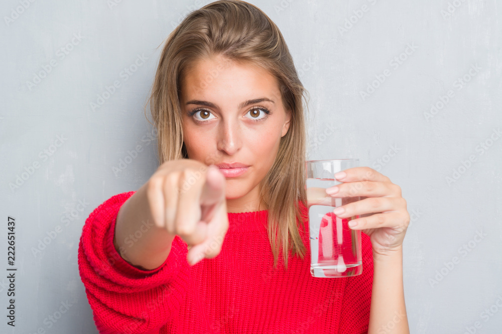 Beautiful young woman over grunge grey wall drinking a glass of water pointing with finger to the camera and to you, hand sign, positive and confident gesture from the front Beautiful young woman over grunge grey wall drinking a glass of water pointing with finger to the camera and to you, hand sign, positive and confident gesture from the front
