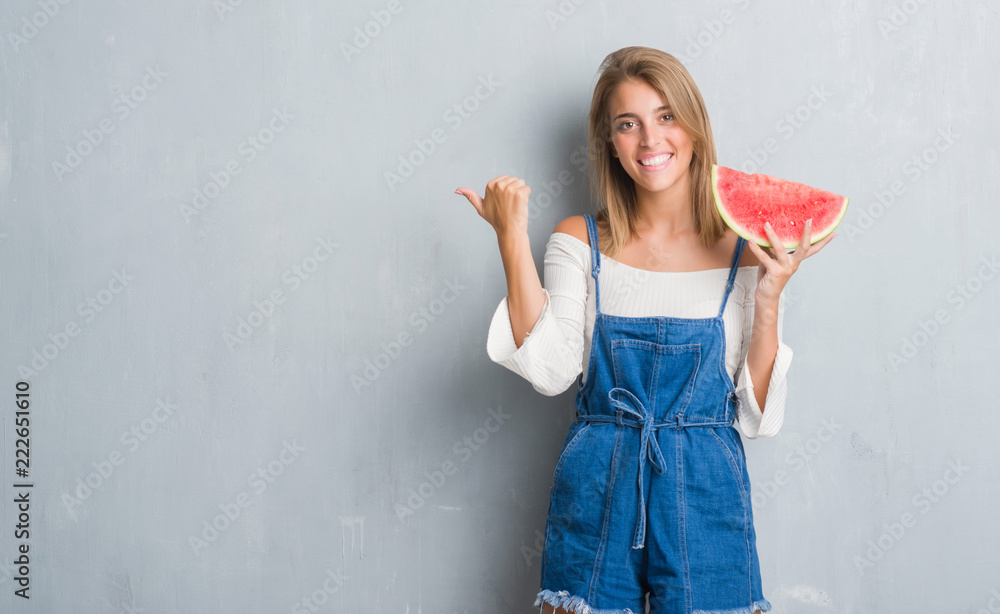 Beautiful young woman over grunge grey wall eating water melon pointing and showing with thumb up to the side with happy face smiling Beautiful young woman over grunge grey wall eating water melon pointing and showing with thumb up to the side with happy face smiling