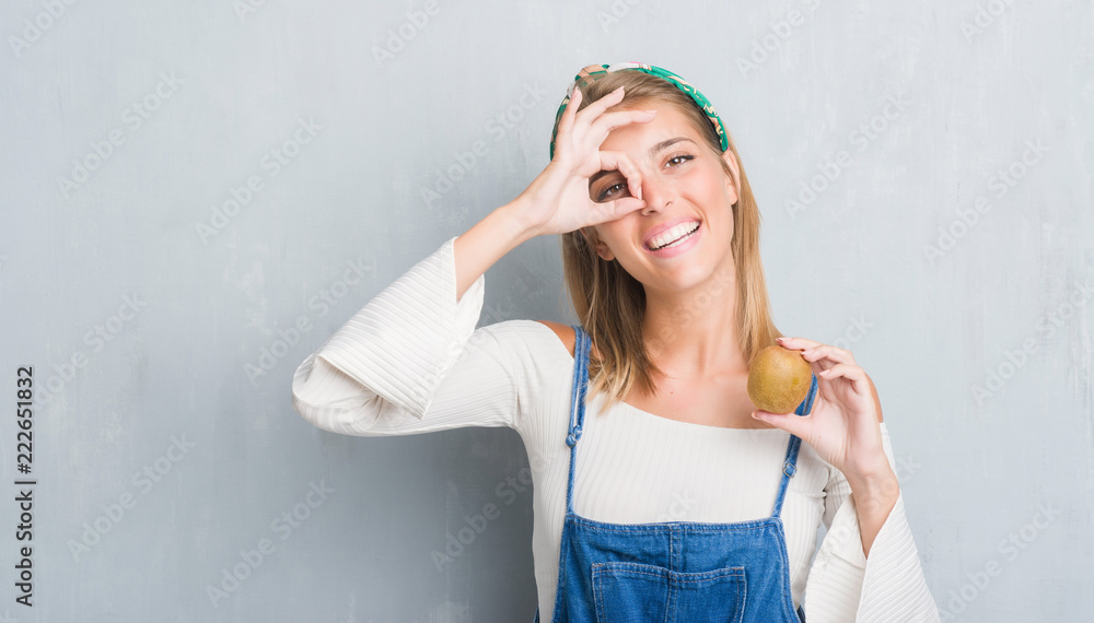 Beautiful young woman over grunge grey wall holding fresh kiwi with happy face smiling doing ok sign with hand on eye looking through fingers Beautiful young woman over grunge grey wall holding fresh kiwi with happy face smiling doing ok sign with hand on eye looking through fingers