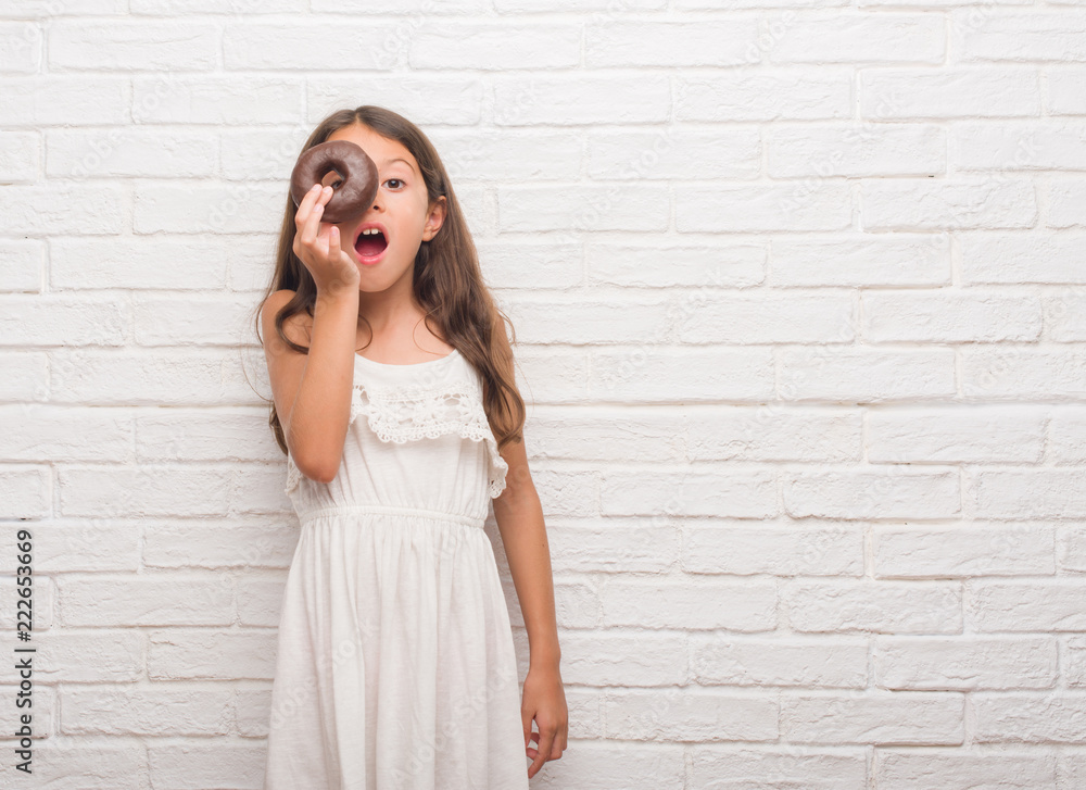 Young hispanic kid over white brick wall eating chocolate donut scared in shock with a surprise face, afraid and excited with fear expression