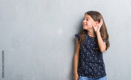 Young hispanic kid over grunge grey wall smiling with hand over ear listening an hearing to rumor or gossip. Deafness concept.