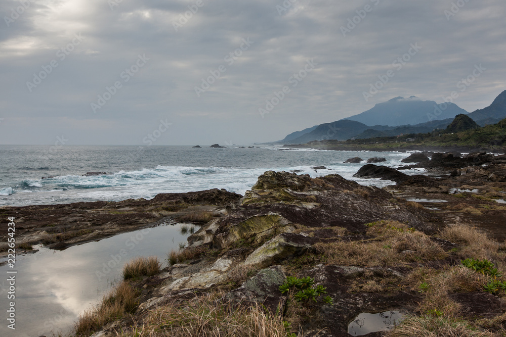 Taiwan East Coast Rocky Coastline Background Image - Overcast Skies, Exotic Rock Formations, Grass and Waves in the Ocean. Ocean Coastline, Asia Landscape Photography, Rays of sunshine in background