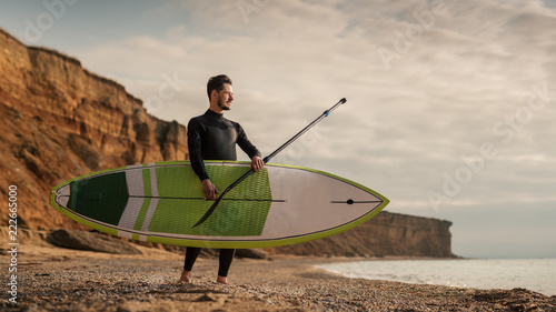 Portrait of a surfer with SUP board on the beach. Young man with a stand up paddleboard at sunset. Extreme sport concept. Male surfer lifestyle. photo