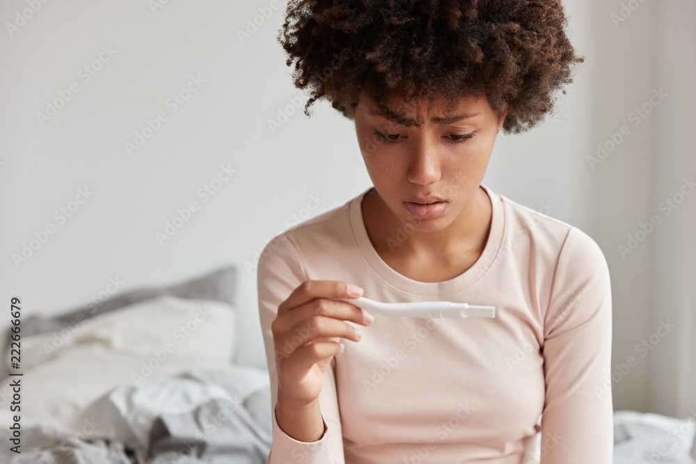 Φωτογραφία Cropped shot of stressful dark skinned woman looks at pregnancy test, doesnt anticipate to notice positive result, isnt ready become mother, sits on bed, dressed in casual clothes