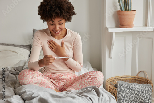 It wil be start of new life! Cheerful African American future mother cant believe in positive result on pregnancy test, awakes early in morning, wears casual pyjamas, poses against bedroom interior photo