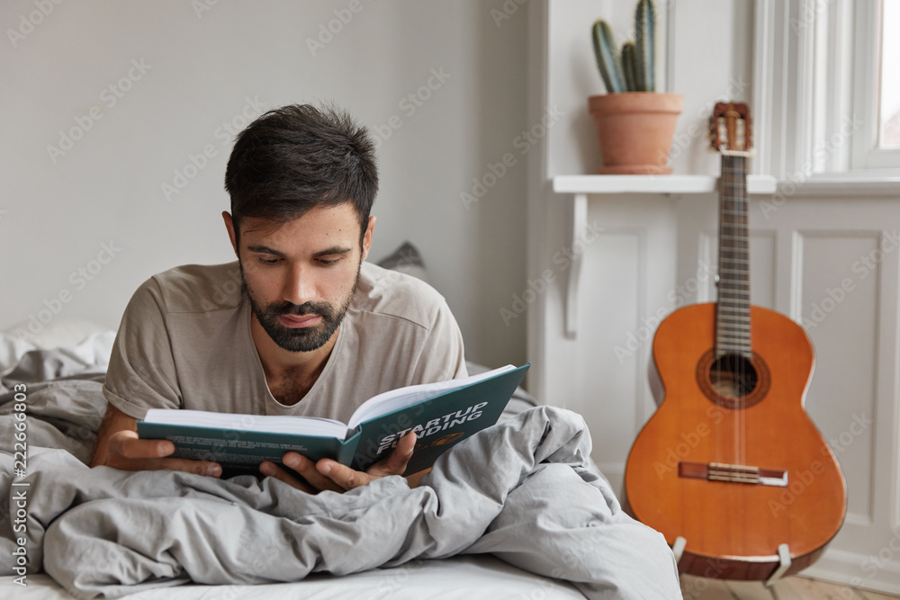 Indoor image of thoughtful bearded guy concentrated on reading, rests in bed. Intelligest student learns information, prepares for exam at home, acoustic guitar in background. Bed time concept