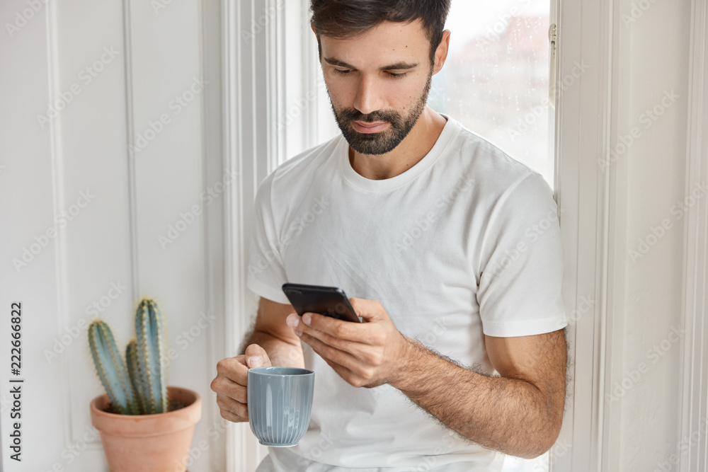Cropped shot of hipster guy in casual white t shirt, uses smart phone for blogging, connected to wireless home internet, drinks fresh coffee, sends text message. Leisure and technology concept Cropped shot of hipster guy in casual white t shirt, uses smart phone for blogging, connected to wireless home internet, drinks fresh coffee, sends text message. Leisure and technology concept