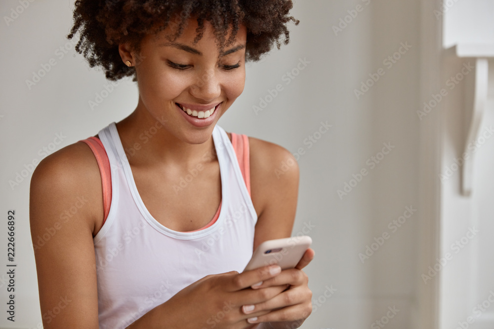 Cropped shot of pleasant looking dark skinned girl makes shopping in popular website, shares news and chats with followers, pays money via online banking service, dressed in casual white vest