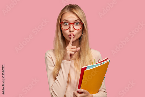Omg, dont speak! Attractive Caucasian girl keeps index finger over lips, demonstrates silence gesture, asks not bother her, be quiet and speechless, wears spectacles with red rim isolated on pink wall