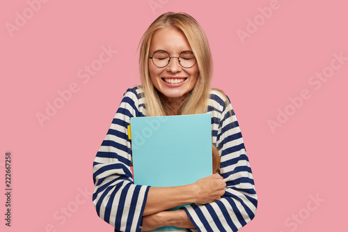 Happy overjoyed smiling teenager with blonde hair, keeps textbook closely, laughs at something funny, has break between classes, poses against pink background, dressed in striped sailor sweater