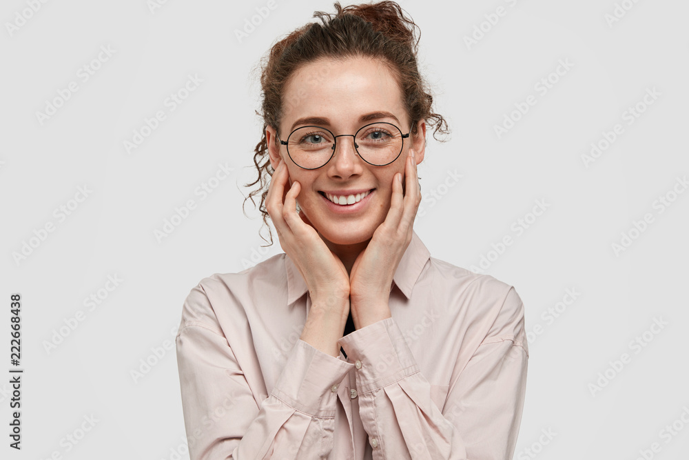Photo of pleasant looking satisfied office worker, dressed in formal clothes, keeps hands on cheeks, has positive expression, being in good mood after successful day, stands against white wall