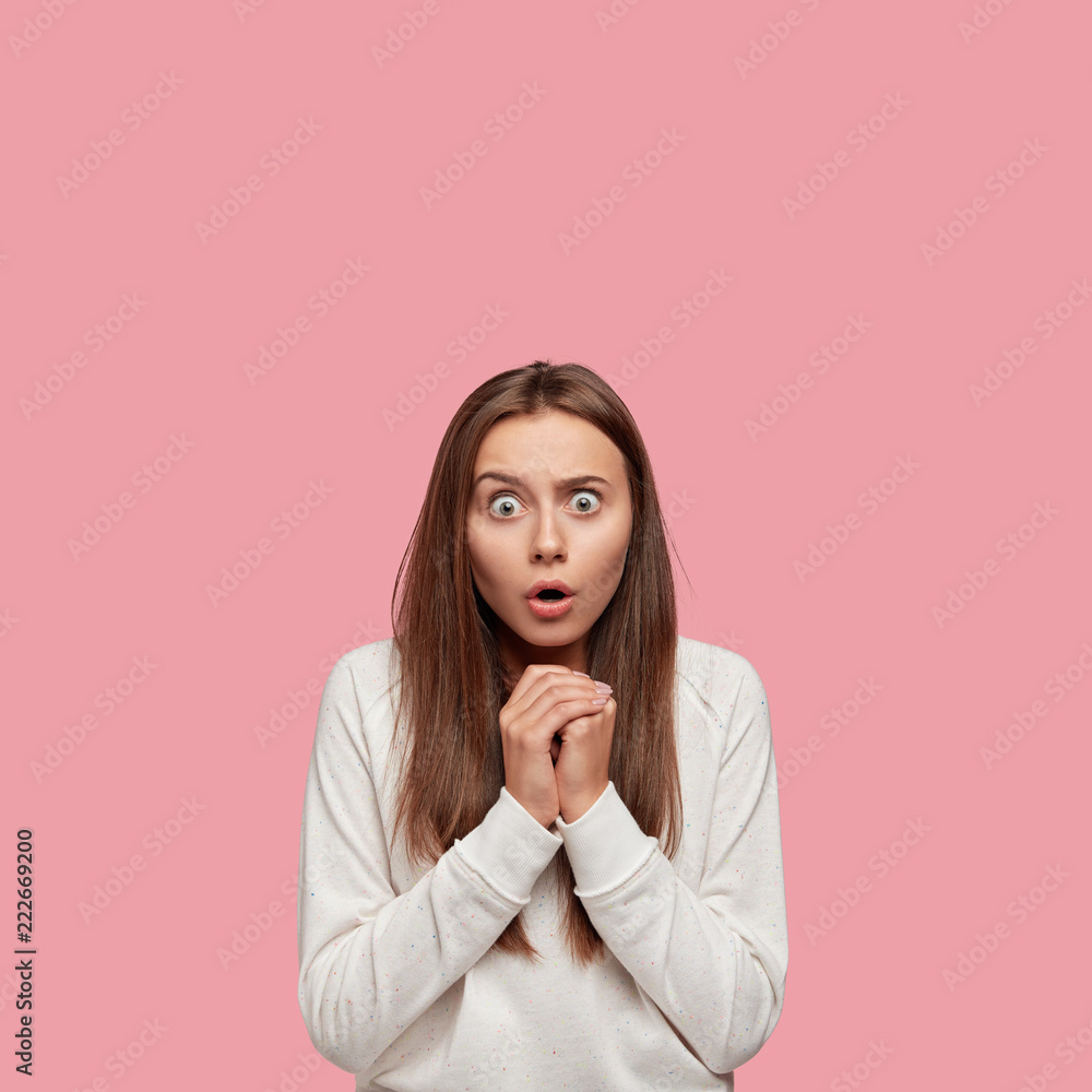 Vertical shot of scared brunette Caucasian woman stares with eyes popped put, keeps hands together, dressed in casual sweatshirt, notices something horrible into distance, isolated on pink wall Vertical shot of scared brunette Caucasian woman stares with eyes popped put, keeps hands together, dressed in casual sweatshirt, notices something horrible into distance, isolated on pink wall
