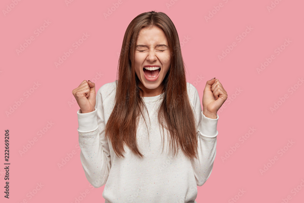 Image of brunette amazed teenager clenches fists with triumph and success, gestures indoor, exclaims with happiness, dressed in casual clothes, isolated over pink studio wall. Body language concept. Image of brunette amazed teenager clenches fists with triumph and success, gestures indoor, exclaims with happiness, dressed in casual clothes, isolated over pink studio wall. Body language concept.