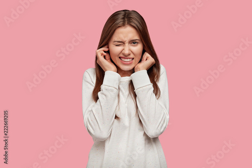 Obraz na płótnie Photo of displeased woman plugs ears with discontent, doesnt want to hear annoying sound or noise, dressed in white sweater, isolated over pink background