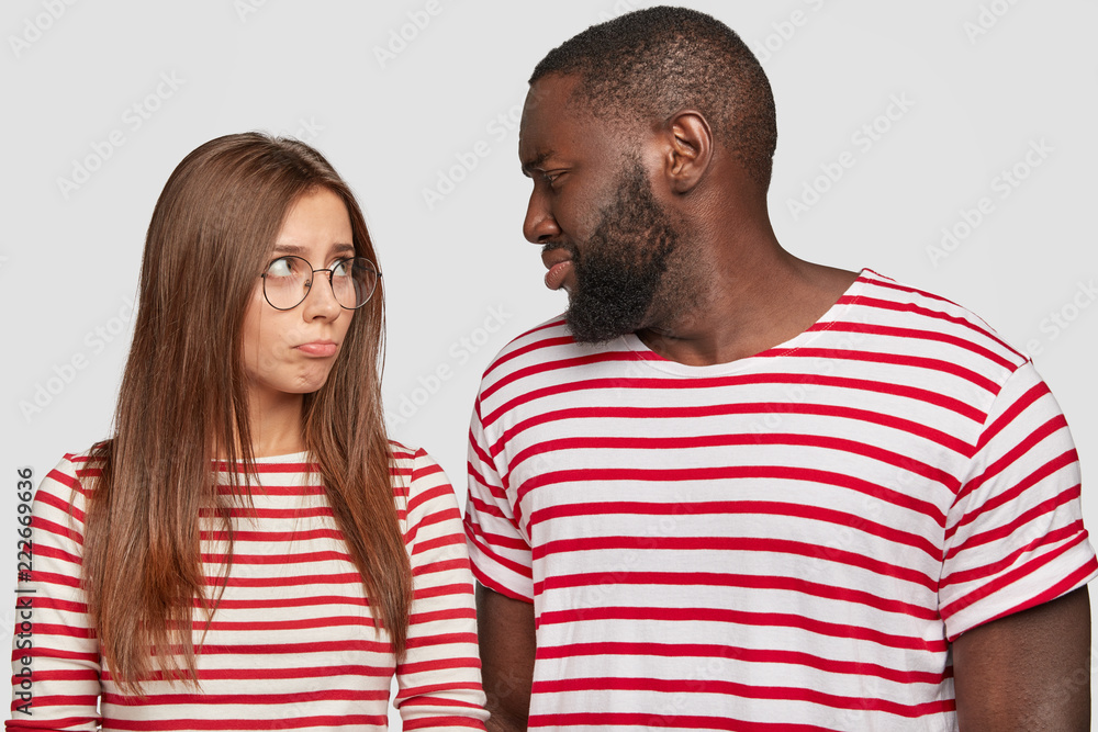Photo of mixed race couple sort out relationships, look in displeasure at each other, have quarrel, dressed in similar t shirts, stand against white background. Interracial family face some problems Photo of mixed race couple sort out relationships, look in displeasure at each other, have quarrel, dressed in similar t shirts, stand against white background. Interracial family face some problems