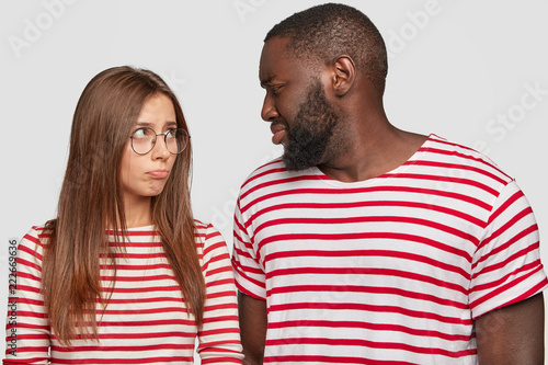 Photo of mixed race couple sort out relationships, look in displeasure at each other, have quarrel, dressed in similar t shirts, stand against white background. Interracial family face some problems