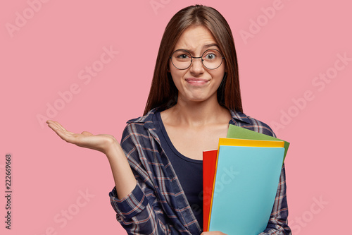 Hesitant doubtful college student holds copy space, frowns face in displeasure, uses textbooks, dressed in checkered casual shirt, isolated over pink background. Puzzled schoolgirl stands indoor
