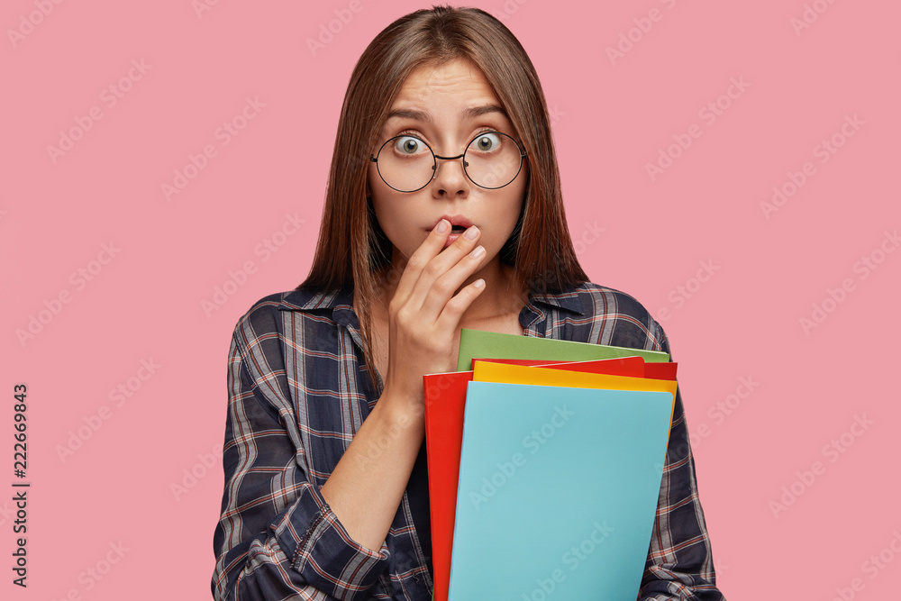 Isolated shot of scared young European woman geek keeps hands on opened mouth, has bugged eyes, dressed in casual clothes, holds colourful folds, dressed casually, isolated over pink background Isolated shot of scared young European woman geek keeps hands on opened mouth, has bugged eyes, dressed in casual clothes, holds colourful folds, dressed casually, isolated over pink background