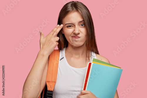 Unimpressed beautiful overworked woman makes hand gun gesture near temple, has displeased facial expression, feels indifferent, tires of constant learning and preparation for classes, stands indoor