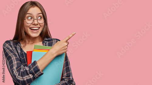 Photo of good looking woman with dark hair, pleasant appearance, indicates with index finger at upper right corner, has good mood after lectures, poses over pink background with blank space.