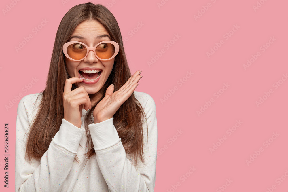 Amazed beautiful young woman with dark hair, wears trendy big shades, smiles broadly at camera, wears casual clothes, poses against pink background with copy space for your advertising content