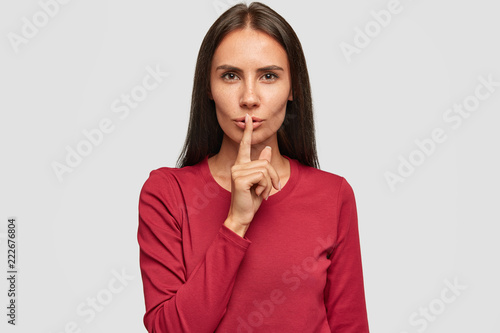 Photo of silent beautiful brunette lady dressed in red sweater, keeps fore finger on lips, has serious expression, stands over white studio background. People, conspiracy and secrecy concept