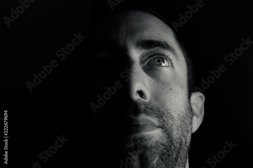 Dramatic portrait of a young man half expressionless face in deep shadow looking up isolated on black background