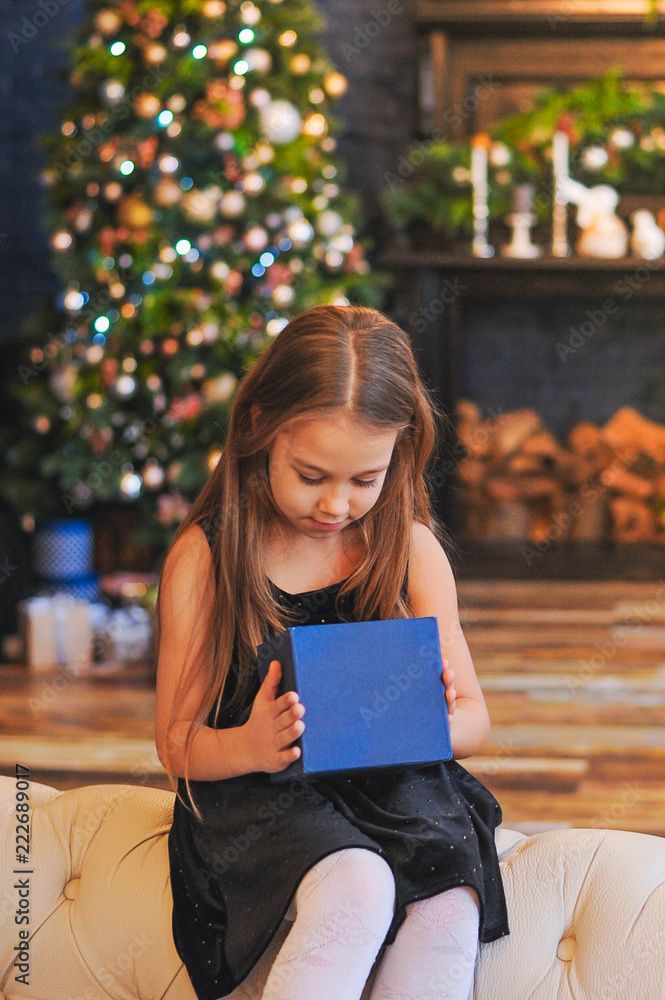 Obraz premium curious little girl smiling while opening Christmas presents. Beautifully decorated Christmas tree and house with lights and lantern.