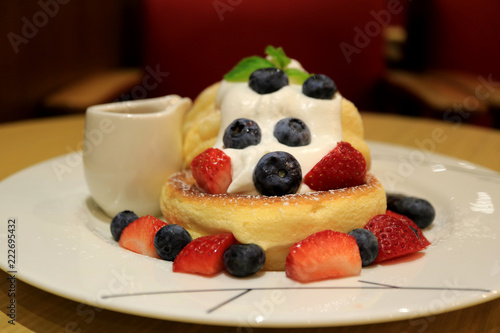Front View of Fluffy Souffle Pancake with Fresh Whipped Cream and Mix Berries on White Plate with Syrup Pitcher 
