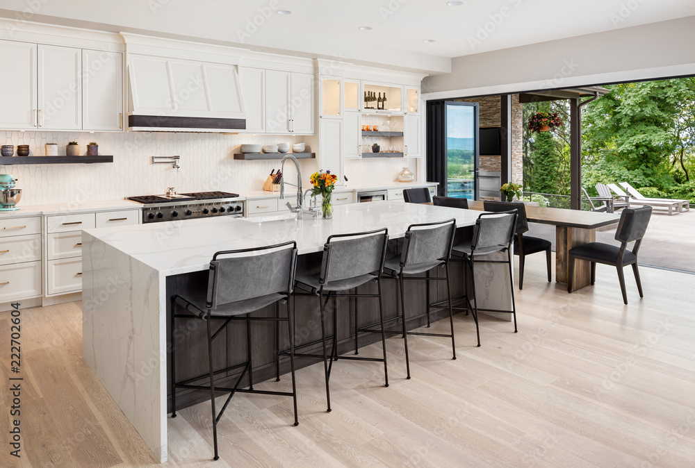 Kitchen interior in new luxury home with hardwood floors, waterfall island, dining room table, and accordion style glass doors. Glass doors are open showing exterior patio