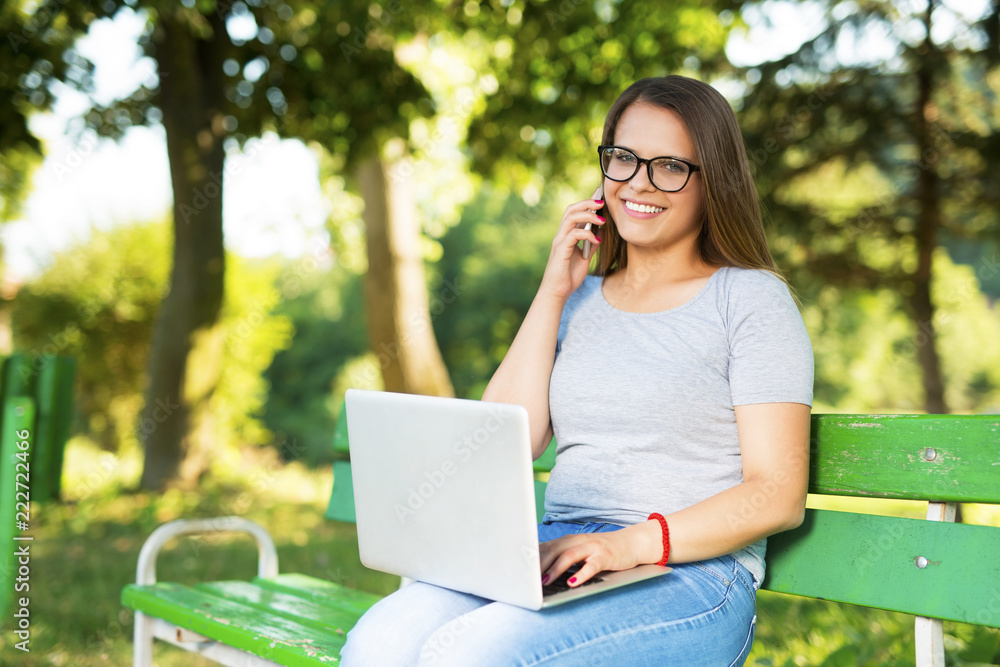Young woman sitting on a bench outdoors in park and using smartphone and laptop Young woman sitting on a bench outdoors in park and using smartphone and laptop