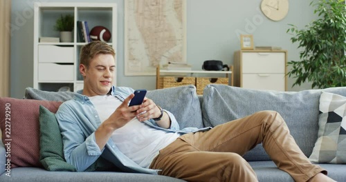 Handsome young man coming in the living room, sitting on the couch and starting chatting on the smartphonr cheerfully. photo