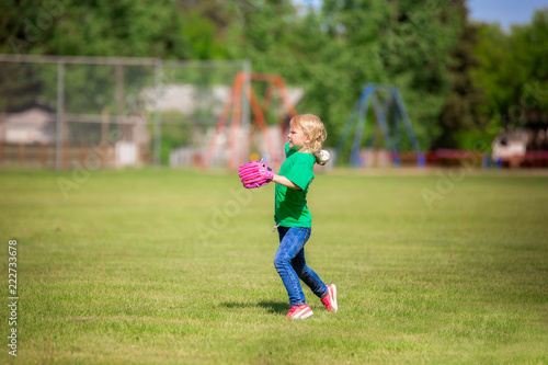 A six year old girl in jeans and t-shirt in action throwing a baseball on green grass on a summer afternoon