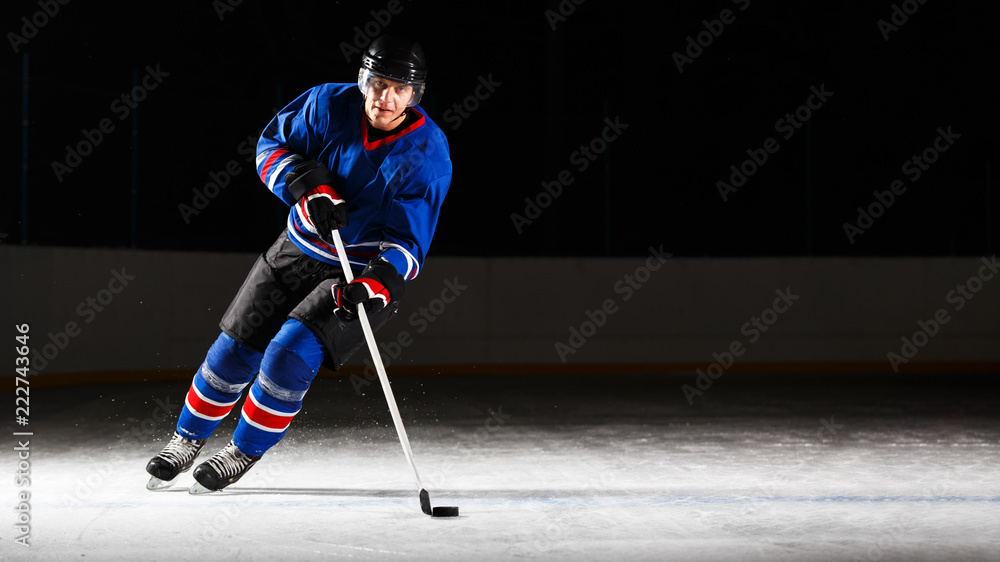 Naklejka premium Young hockey player with stick and puck skating on rink in attack against dark background