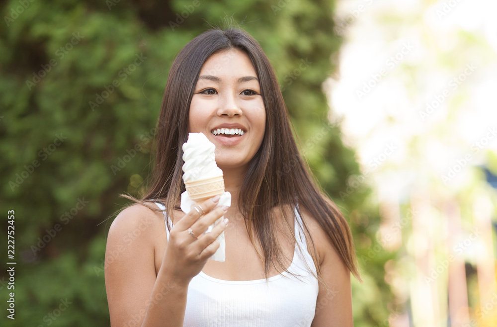 Happy young smiling asian brunette woman eating ice cream on green trees background in Park