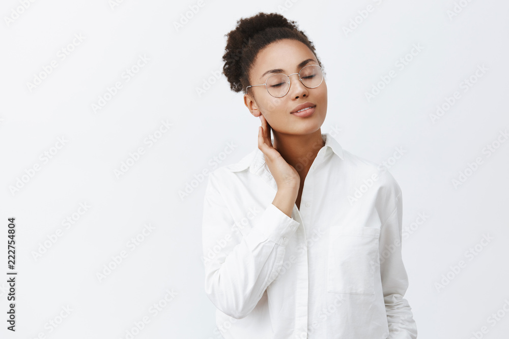 Girl recalls passionate kiss of lover. Portrait of tender feminine african-american woman in glasses and white shirt, closing eyes and touching neck gently, standing over grey background