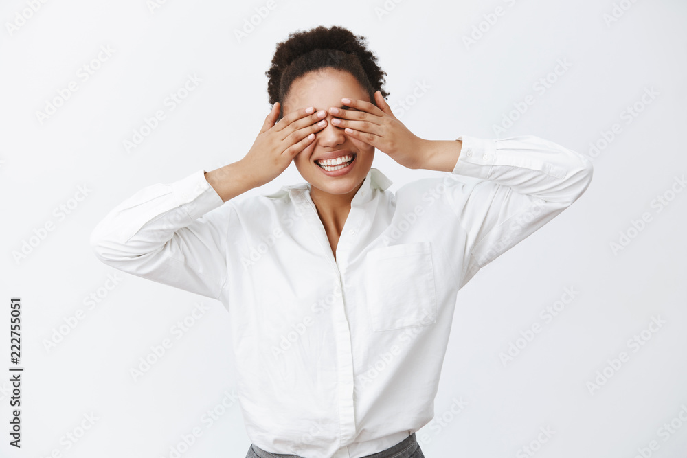 Indoor shot of charming carefree african-american female employer with white shirt, covering eyes with palms, being blinded while playing peek-a-boo and smiling broadly at camera over grey wall Indoor shot of charming carefree african-american female employer with white shirt, covering eyes with palms, being blinded while playing peek-a-boo and smiling broadly at camera over grey wall
