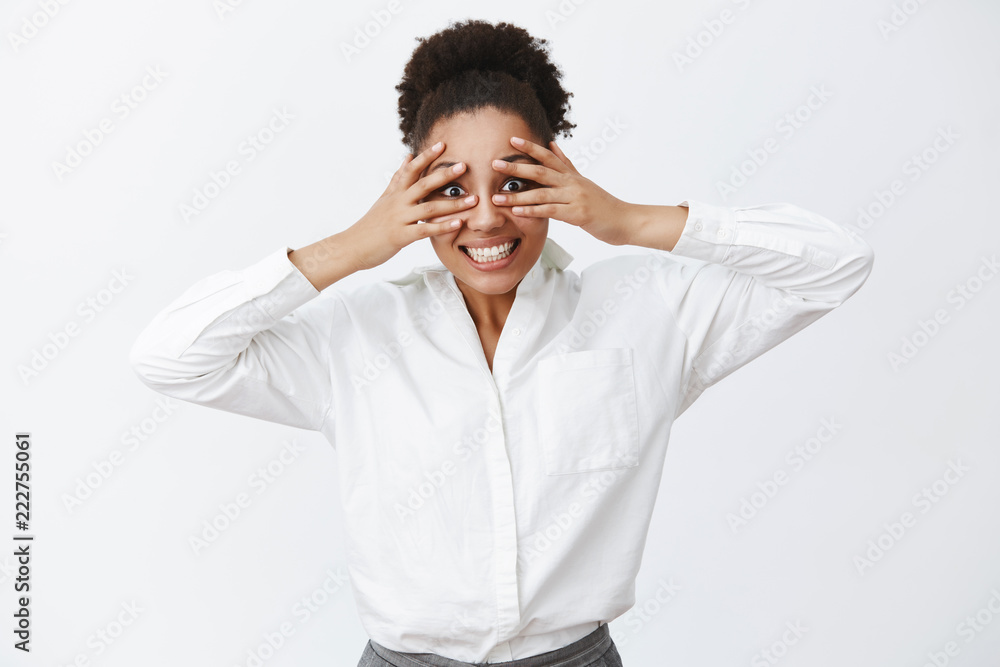 Looking right at you. Portrait of charming carefree active businesswoman with dark skin in white office shirt, covering eyes with palms and peeking through fingers, making funny face, smiling Looking right at you. Portrait of charming carefree active businesswoman with dark skin in white office shirt, covering eyes with palms and peeking through fingers, making funny face, smiling