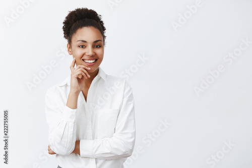Good-looking successful dark-skinned female entrepreneur listening with polite and friendly smile employee, applying for new job, interviewing person, holding finger on cheek and smiling broadly