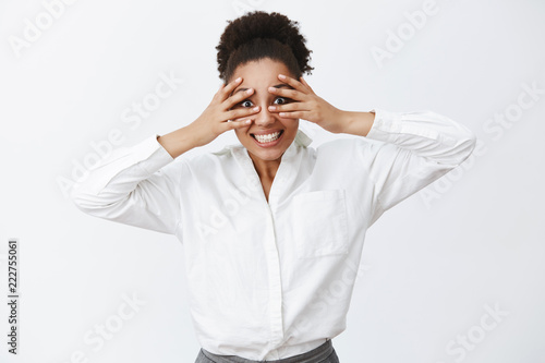 Looking right at you. Portrait of charming carefree active businesswoman with dark skin in white office shirt, covering eyes with palms and peeking through fingers, making funny face, smiling