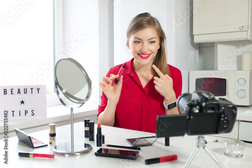 Indoor shot of young European Russian woman making video tutorial about applying makeup being good expert in beauty industry, feeling confident and smiling positively at camera standing on table