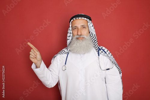 Canvas Print Arabic old bearded hospital doctor with stethoscope posing on red background