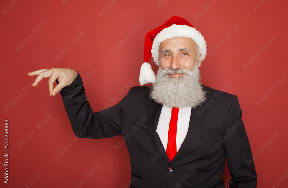 Senior man in santa hat an suit presenting something on a red wall, smiling for the camera. Xmas concept. Senior man in santa hat an suit presenting something on a red wall, smiling for the camera. Xmas concept.
