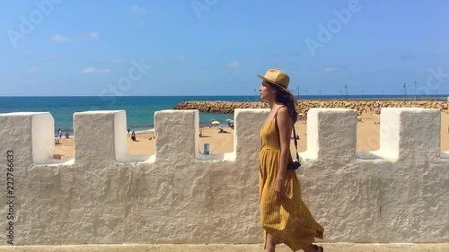 Tourist or casual girl in beautiful yellow dress walking along the wall next to the Asilah beach on her vacations. Morocco time. 4K