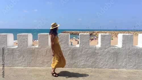 Tourist or casual girl in beautiful yellow dress walking along the wall next to the Asilah beach on her vacations. Morocco time. 4K