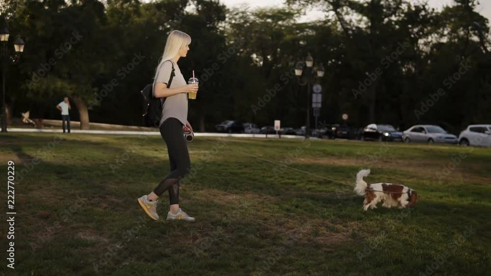 Young beautiful woman with long blonde hair walking with her dog in the park .She is wearing casual clothes and holding drink in hand Young beautiful woman with long blonde hair walking with her dog in the park .She is wearing casual clothes and holding drink in hand