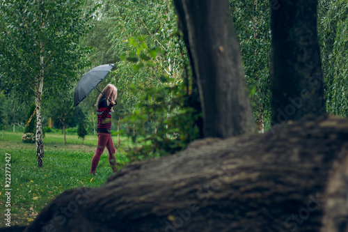 creative foreshortening between trees of lonely teenager girl walk in autumn park natural place under umbrella in wet rainy gray day time 