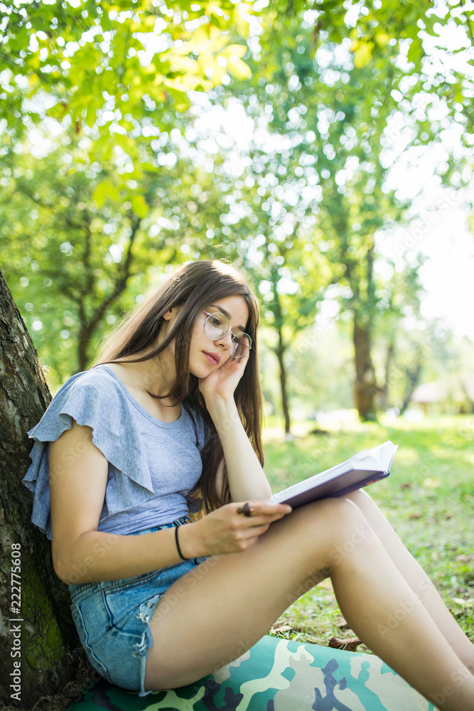 Young woman sitting and reading her favorite book on a o green gras under tree in a nice sunny summer Young woman sitting and reading her favorite book on a o green gras under tree in a nice sunny summer