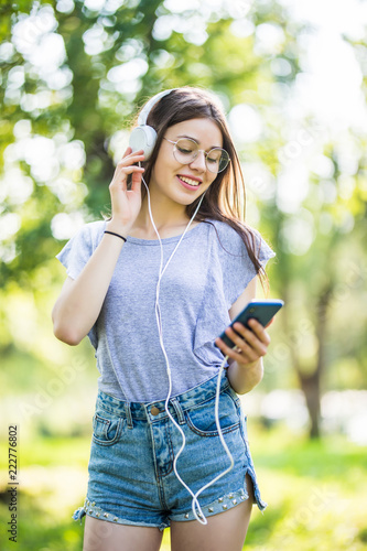 Smiling young girl student holding mobile phone, walking at the park, listening to music with earphones